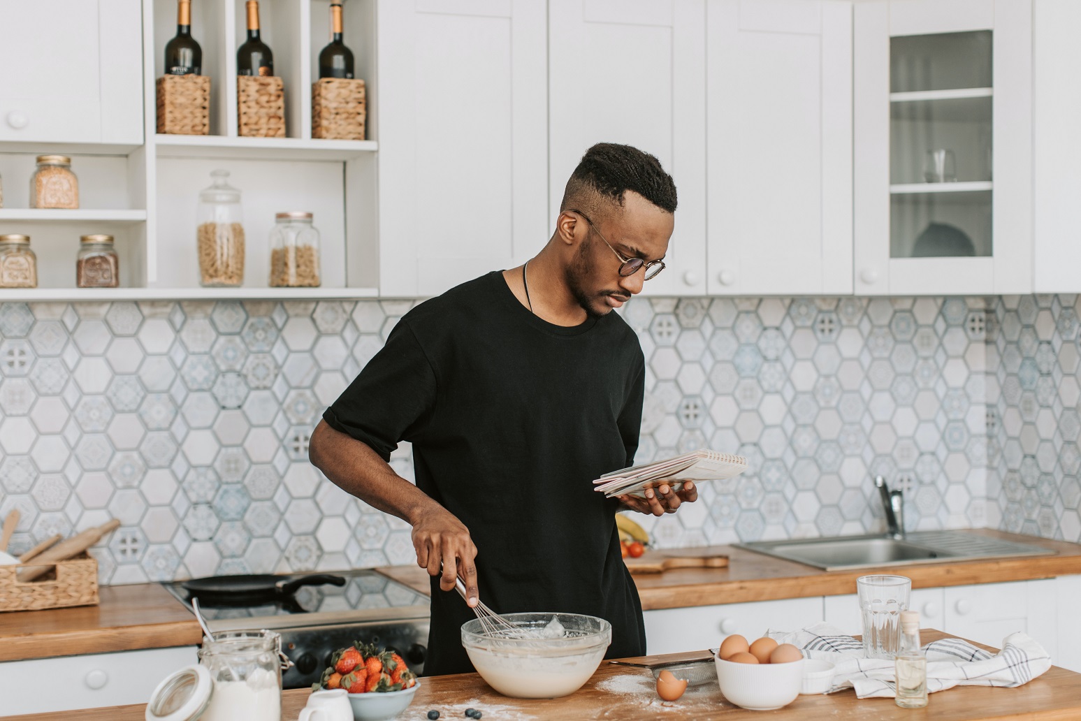 Photo of a man in a kitchen, looking at a notebook while mixing something in a bowl. The bowl is surrounded by eggs, strawberries and other ingredients.