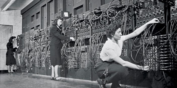 Black and white photo of three women plugging in cables along a big wall of cables and switches (the ENIAC computer)