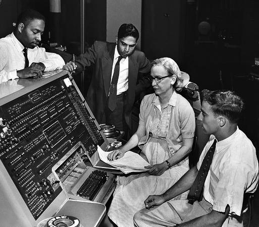 Photo of Grace Hopper seated at a big board of switches (a computer), with several men around her