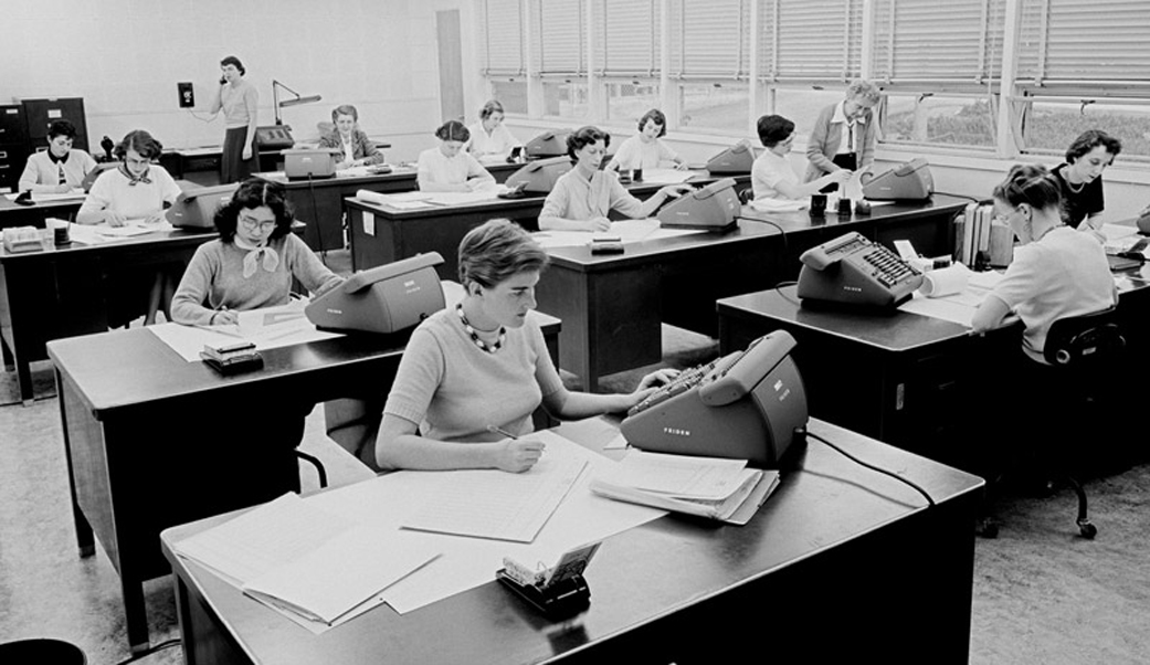 Black and white photo of rows of desks. At each desk is a woman with a pile of papers and some sort of large machine with buttons on it, perhaps a calculator.